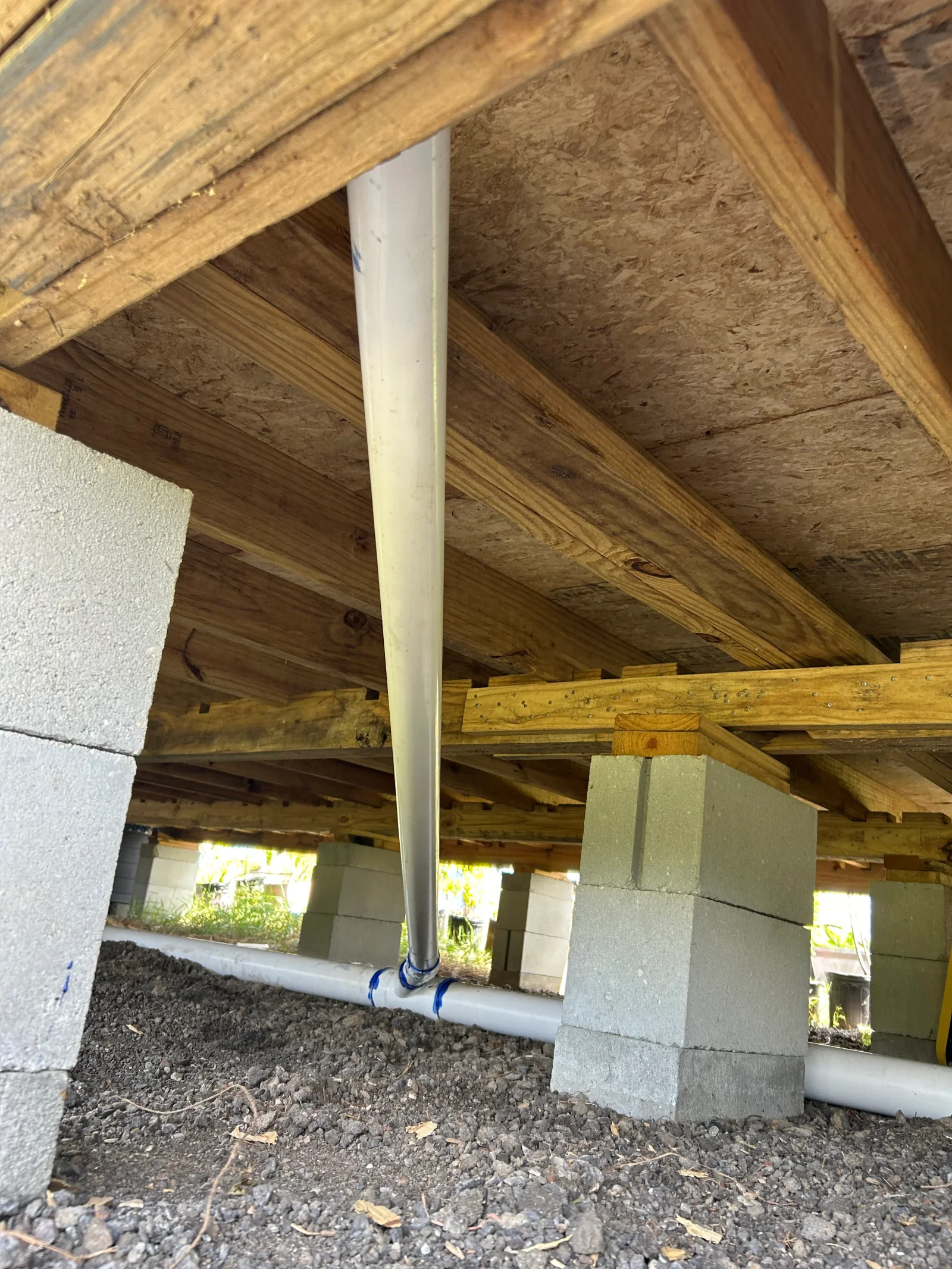 White PVC drain pipe with blue-taped joint running under wood-framed floor on concrete block piers over gravel crawlspace