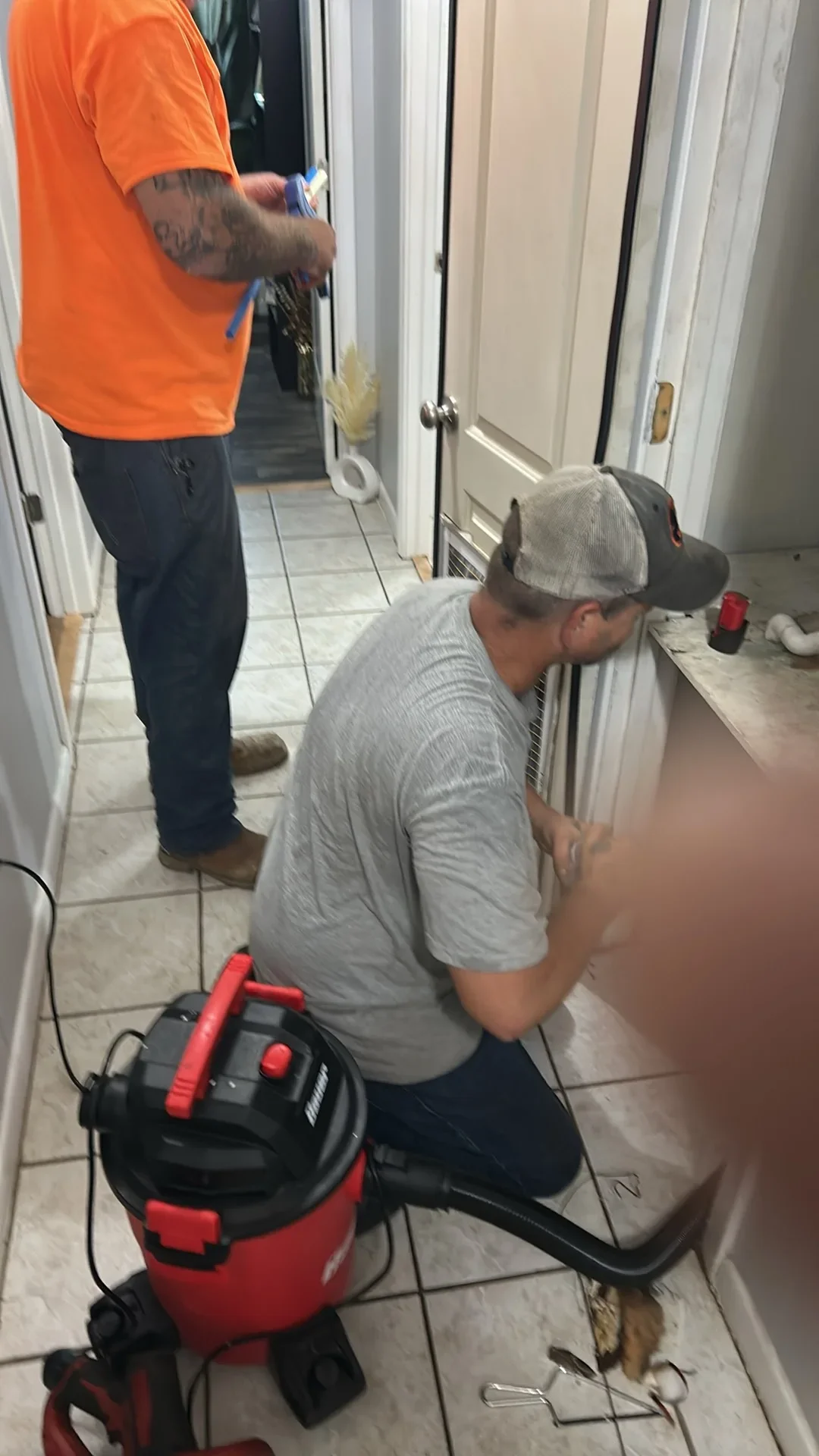 Technician vacuuming debris from a floor vent with a red wet/dry vac in a tiled hallway, white PVC pipe visible