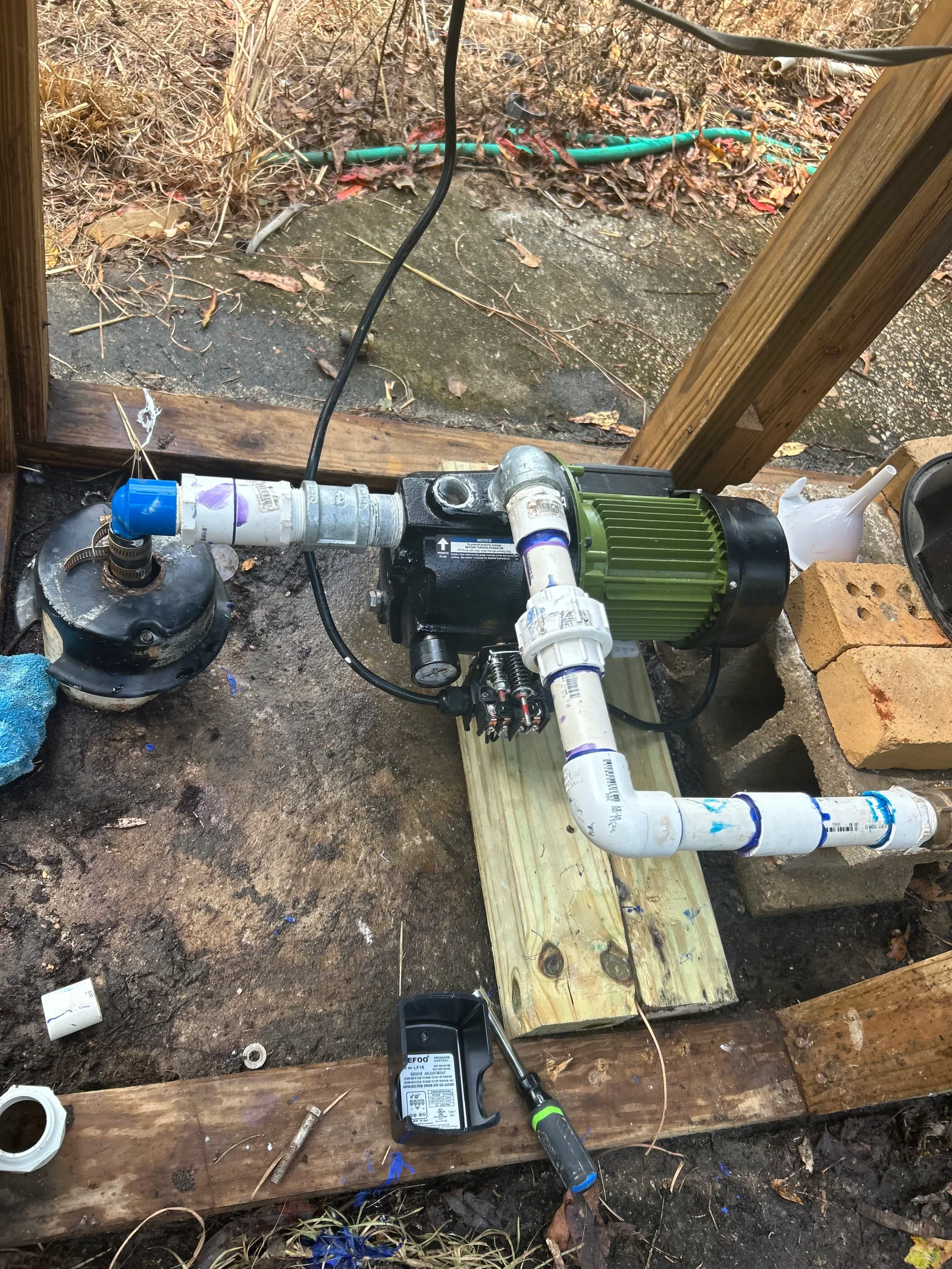 Jet well pump with green motor, black pressure tank, white PVC piping, and pressure switch on wood base