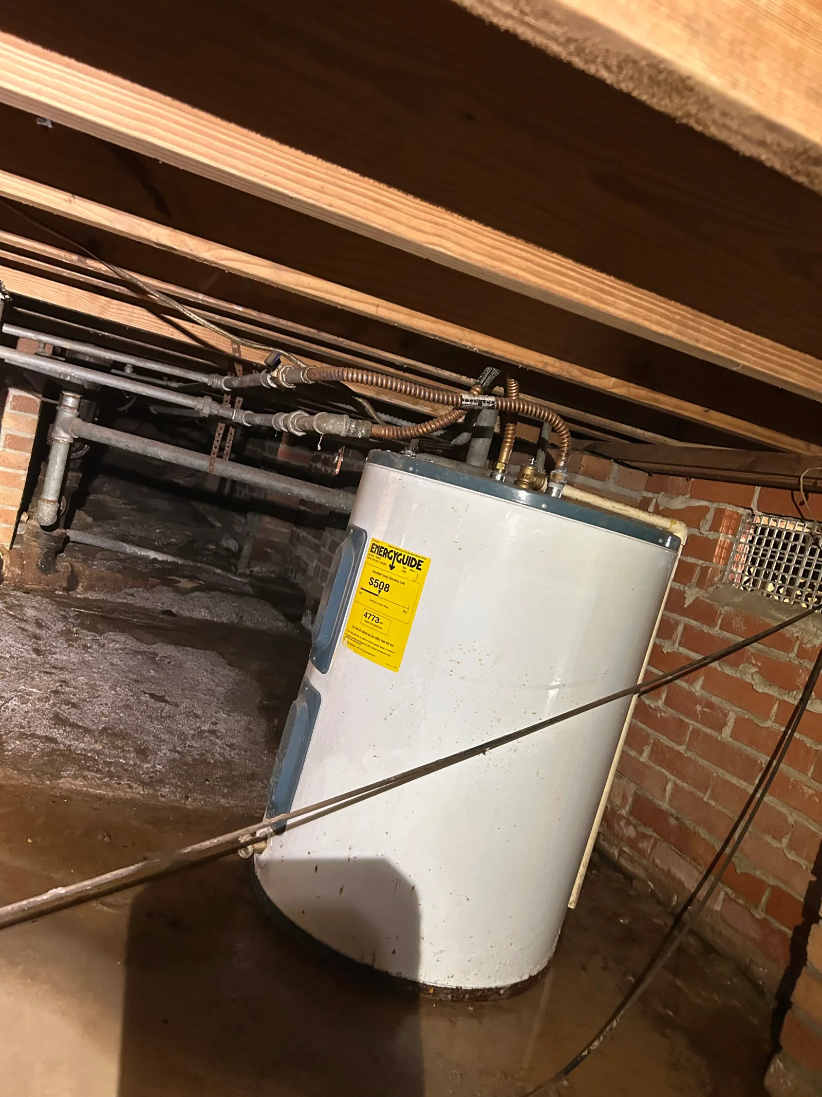 White gas water heater with copper connectors and galvanized pipes in a crawl space under wood joists