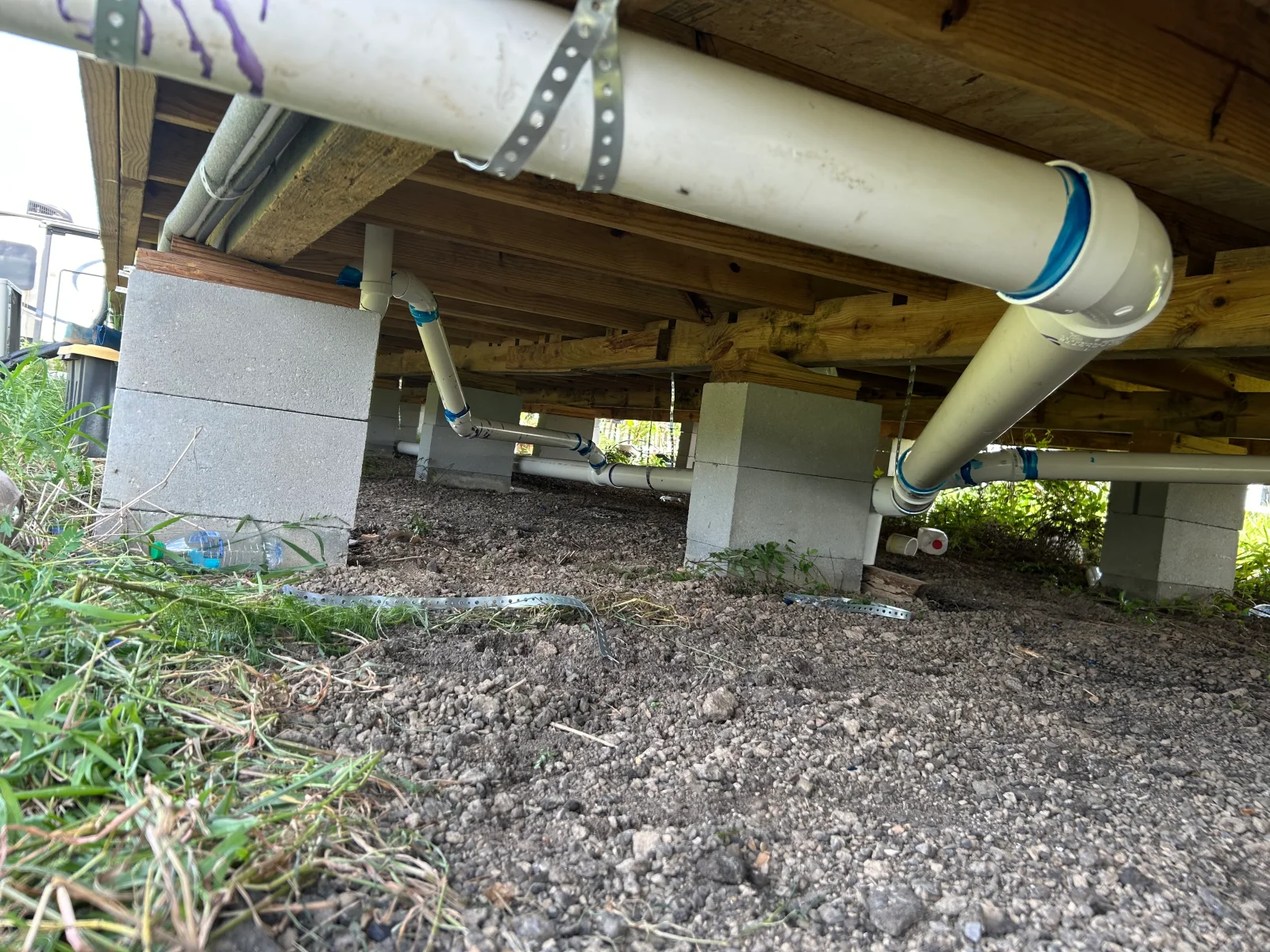 Underfloor PVC drain lines with blue-banded fittings, metal strap hangers, and concrete block piers in a crawlspace